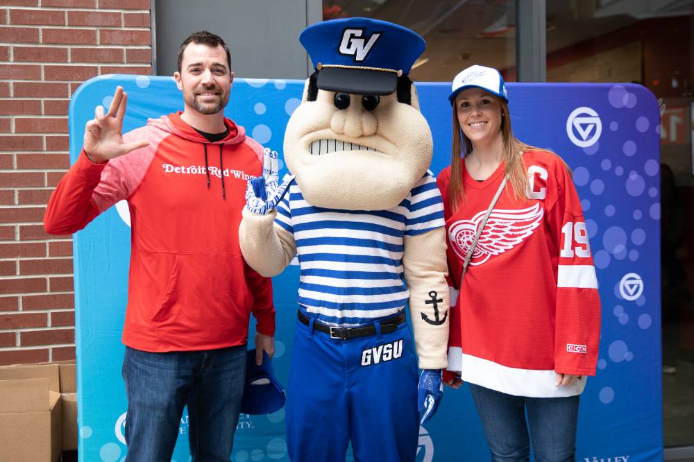 Two alumni pose with Louie while doing the Laker hand gesture at the Detroit Red Wings GVSU Night